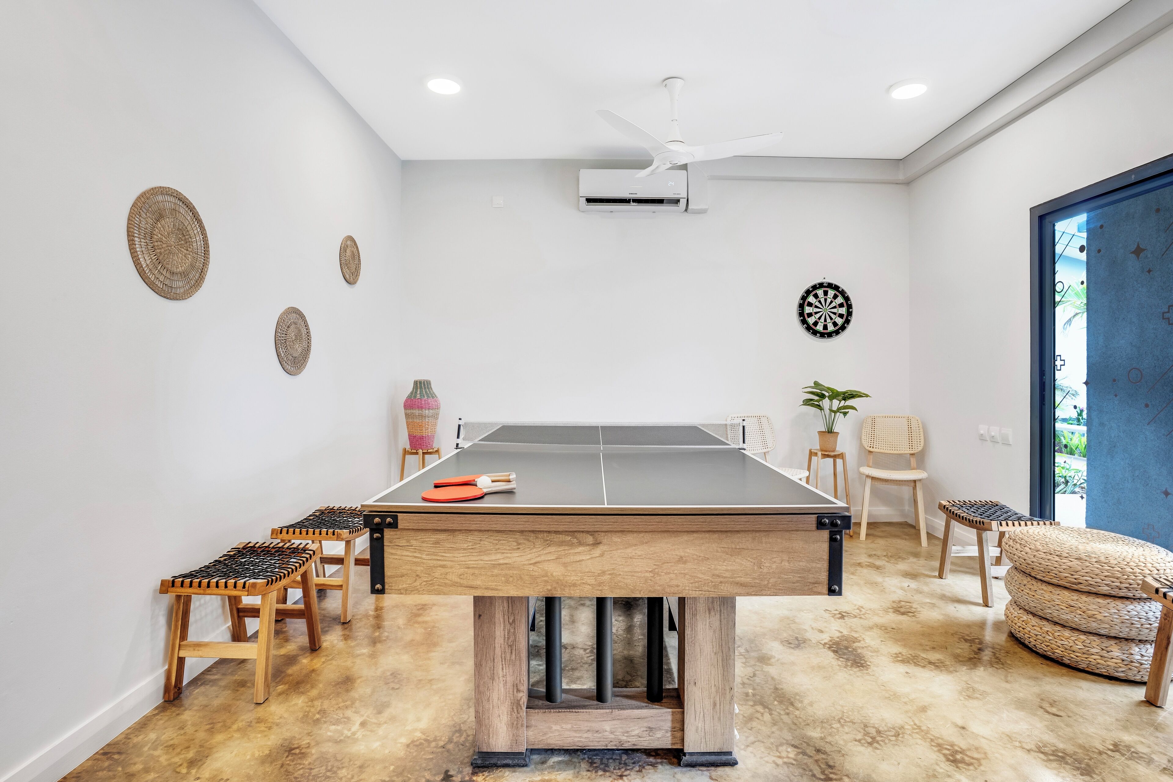 Una mesa de ping pong con taburetes, un ventilador de techo y un dardo en una habitación con piso de concreto.