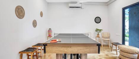 Una mesa de ping pong con taburetes, un ventilador de techo y un dardo en una habitación con piso de concreto.