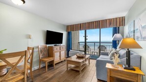 A modern living room with a wooden floor, a flat-screen TV, a sofa, a dining table, and a balcony with a view.