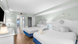 A hotel room with two beds, a desk, and a lamp. There is a view of the sea through an open door.