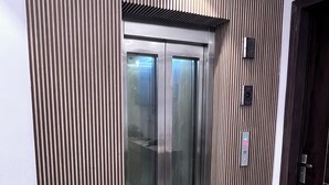 An elevator with a metallic finish and glass doors, flanked by vertical wooden panels. - HAVELIYA HOMES BB RESIDENCY (New Delhi)
