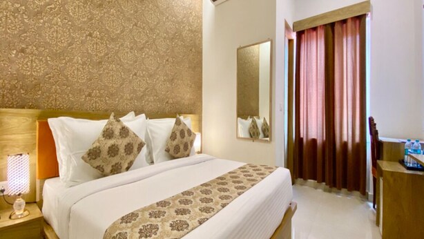 A hotel room with a large bed, a nightstand, an air conditioning unit, a mirror, a desk with a chair, and a window with curtains. - HAVELIYA HOMES BB RESIDENCY (New Delhi)