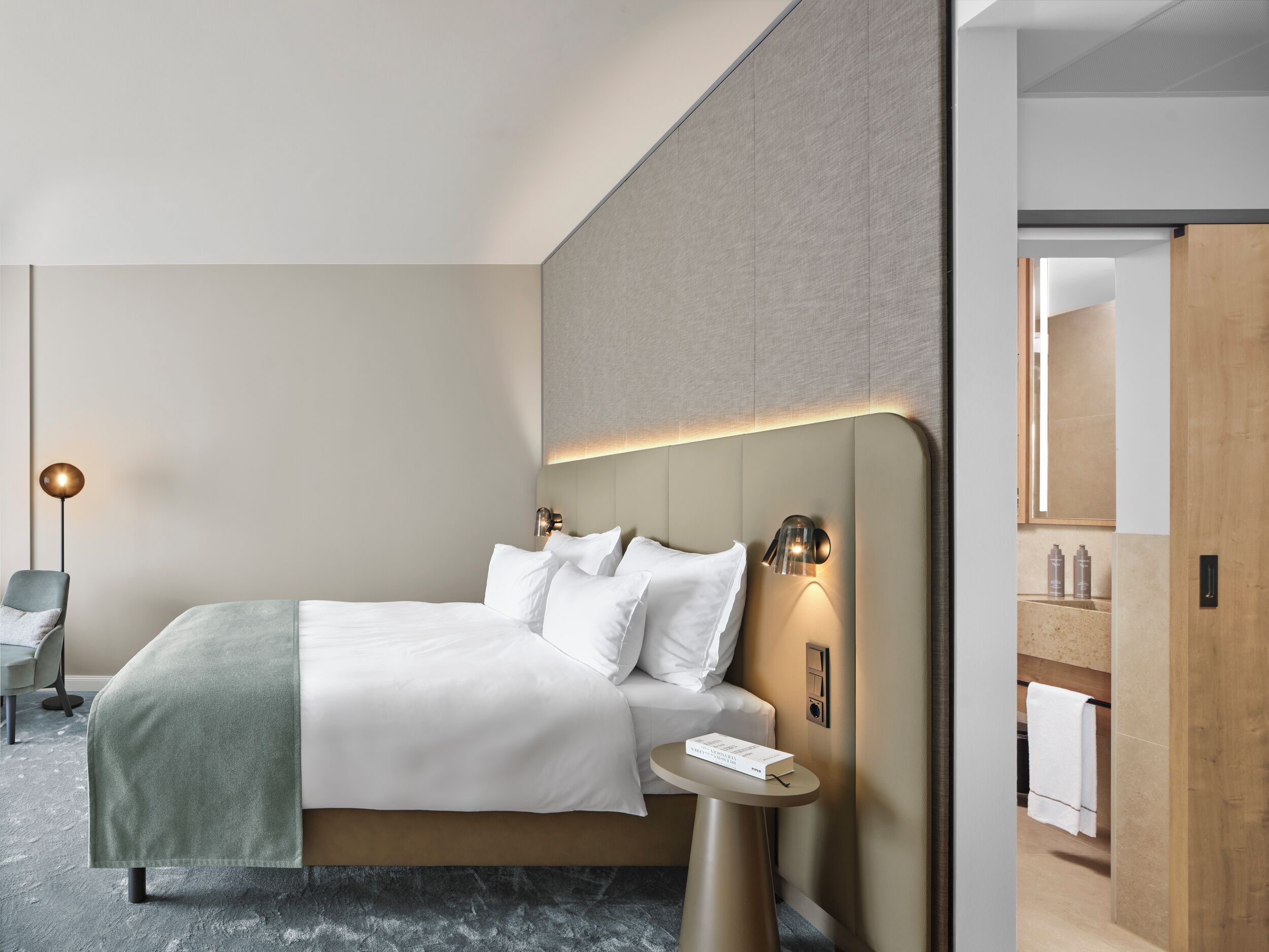 A modern hotel room with a large bed, bedside tables, a wall-mounted light, and a view into the bathroom.