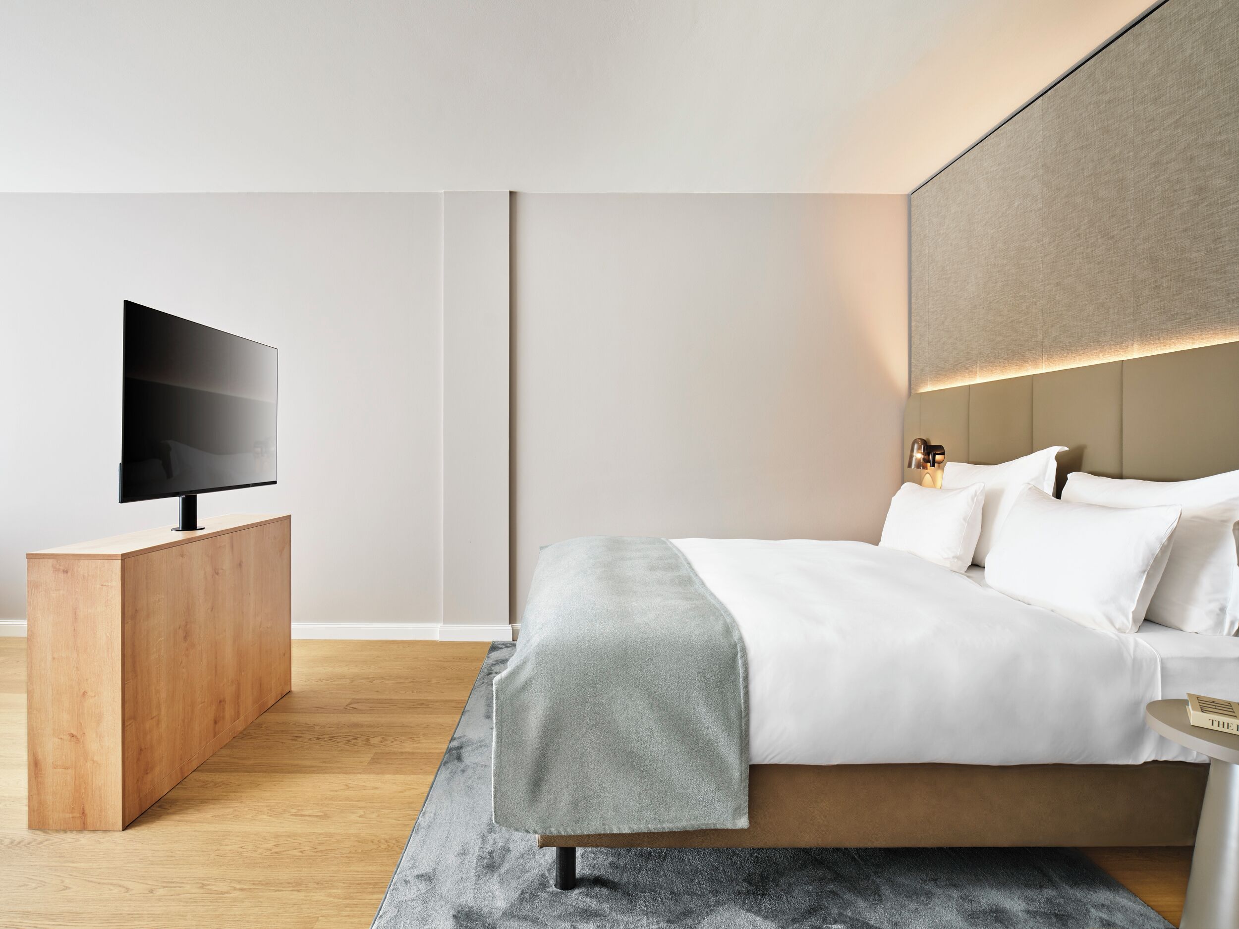 A modern hotel room with a flat-screen TV on a wooden console, a large bed with white linens, and a grey throw blanket.