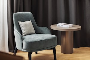 Un sillón gris con un cojín, una mesa redonda con un libro titulado 'LOEWE', y un telón oscuro de fondo.