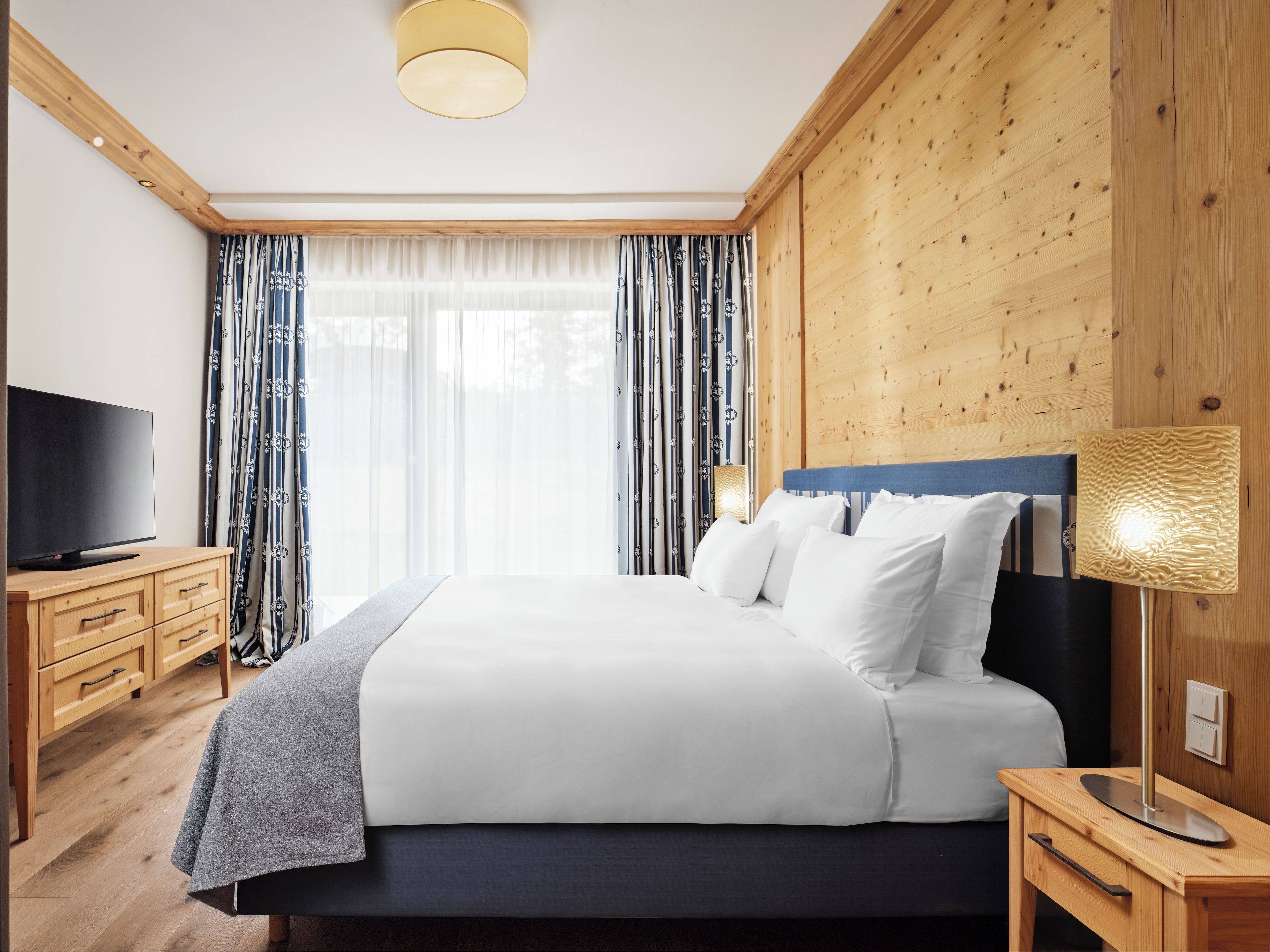 Una habitación de hotel moderna con una cama grande, cabecera de madera, un televisor de pantalla plana y una mesita de noche con lámpara.