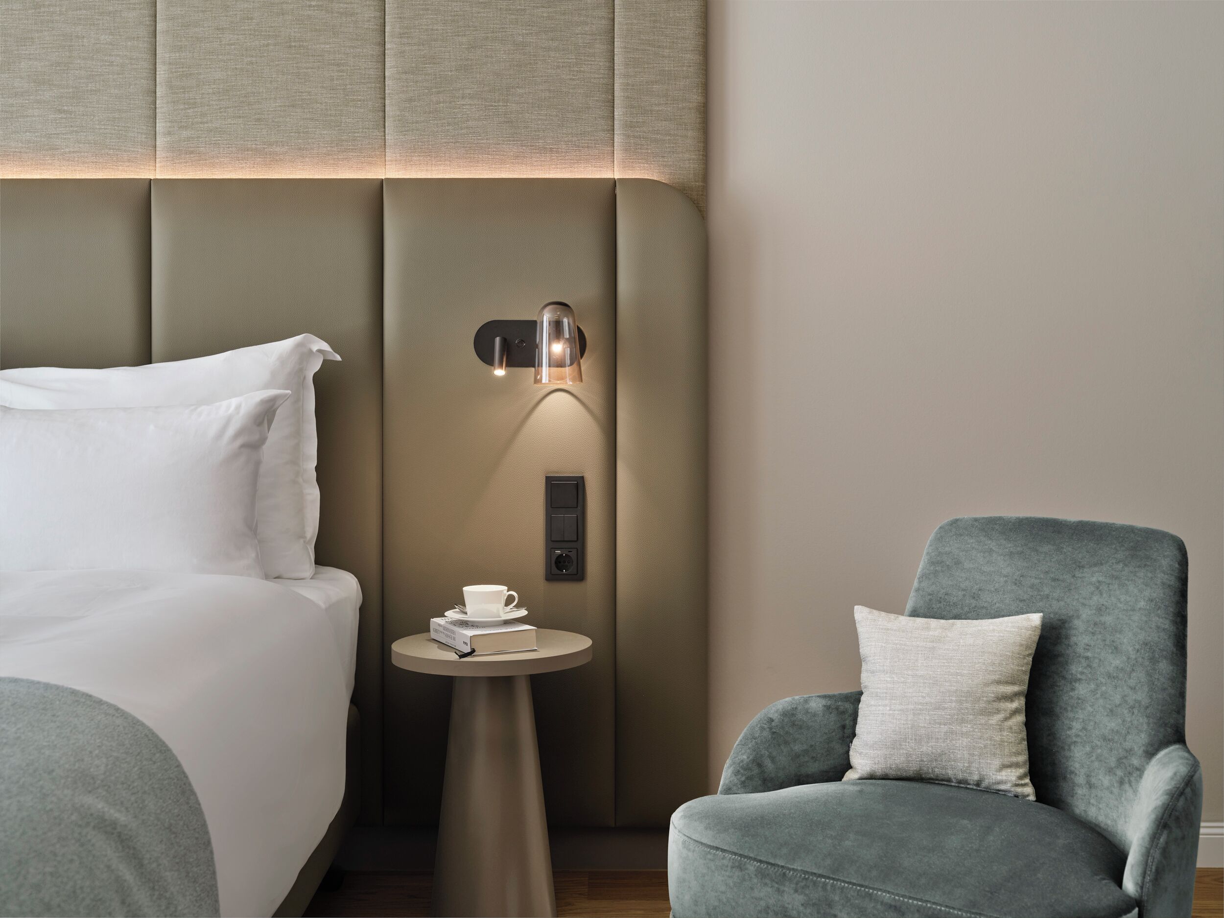 Ein Hotelzimmer mit einem Bett, einem kleinen runden Tisch mit Tasse und Buch, einem Sessel und einer Wandleuchte.