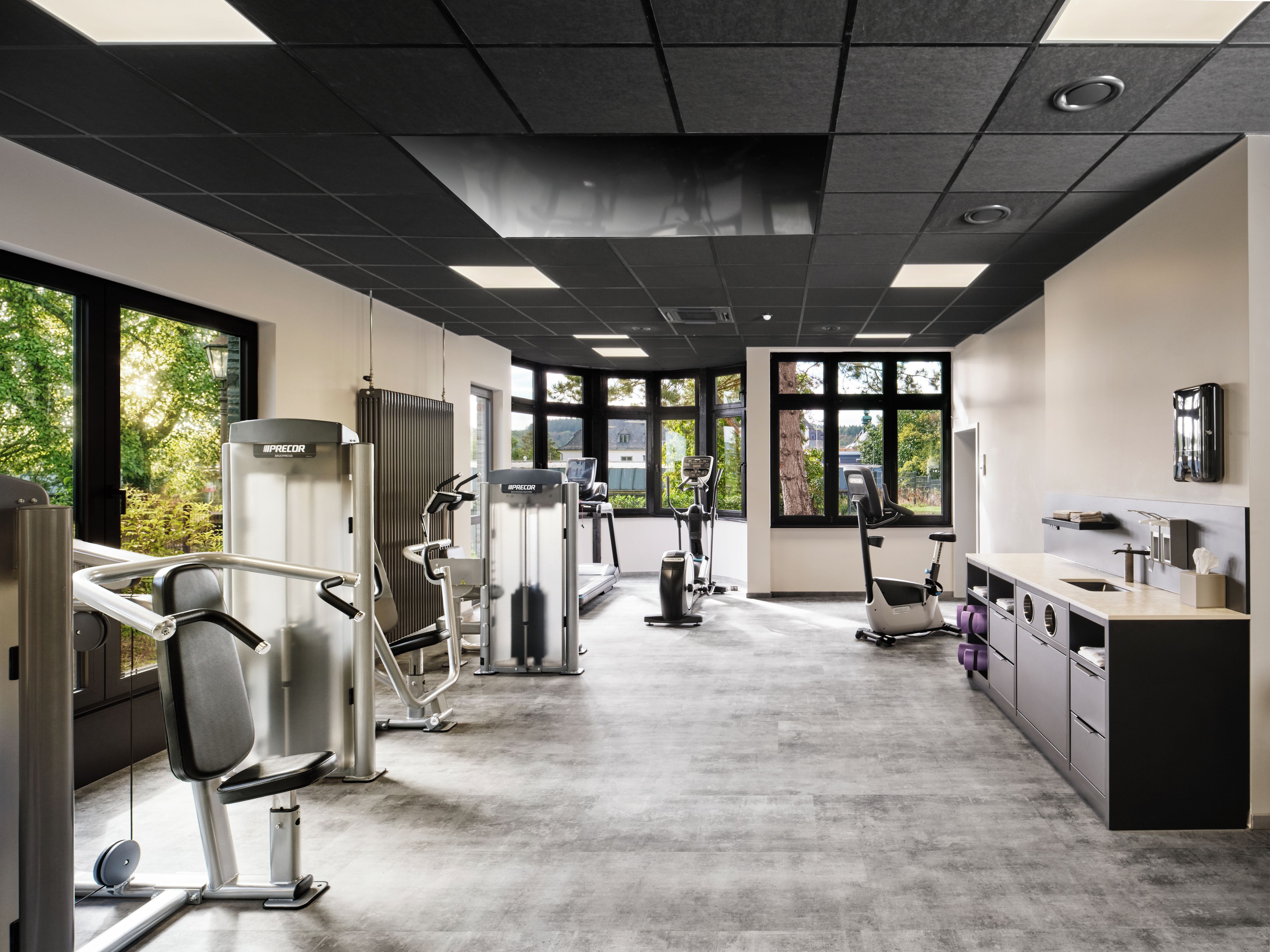 A gym with various exercise machines, large windows, and a kitchenette area.