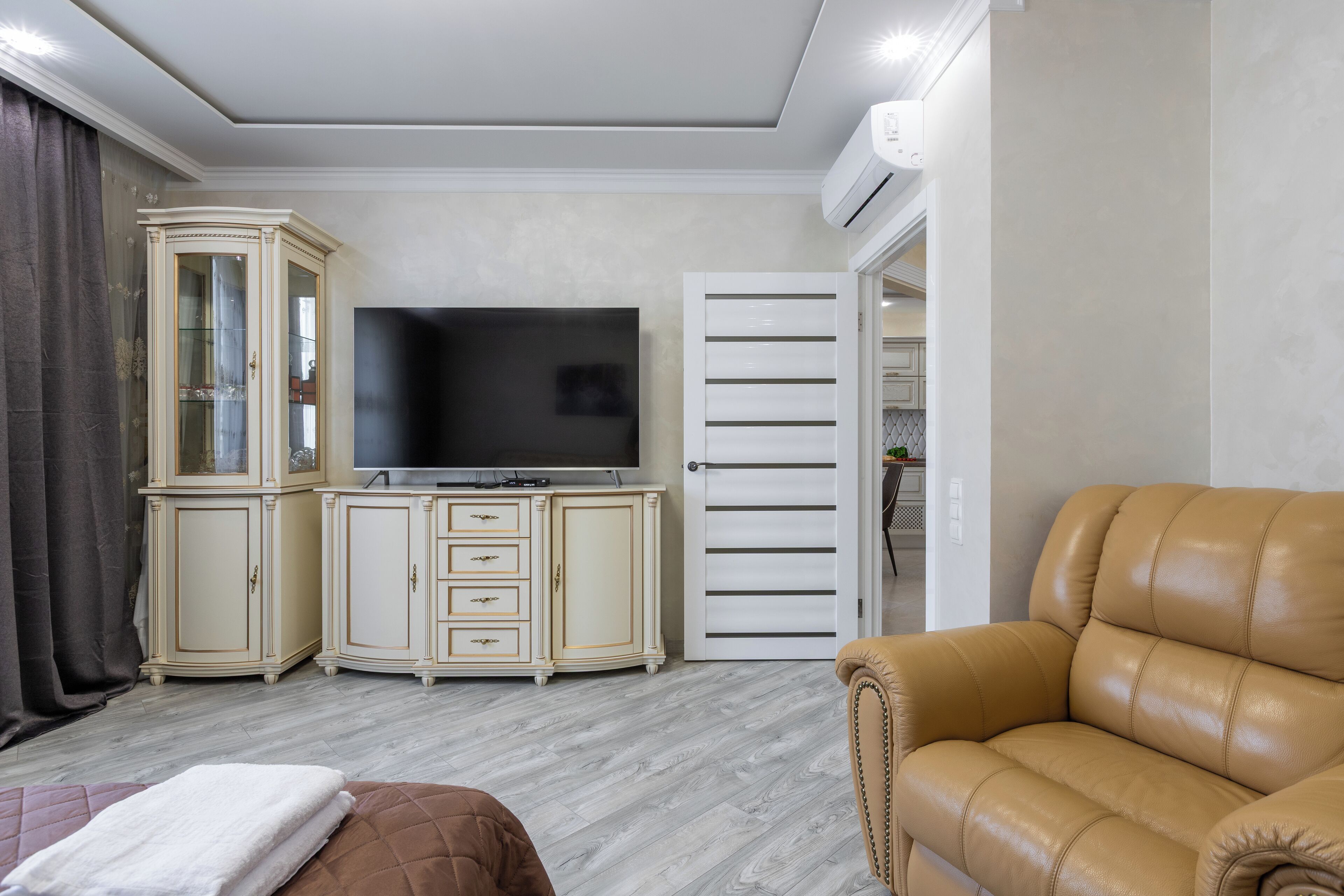 Ein Wohnzimmer mit Flachbildfernseher, einem beige-farbenen Ledersofa, einer Vitrine und einer weißen Tür.