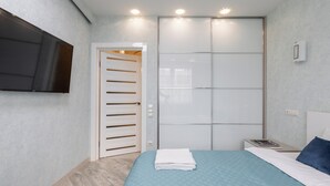 Ein Schlafzimmer mit einem Bett, einem an der Wand montierten Fernseher, einem Kleiderschrank und einem Nachttisch mit Buch und Lampe.