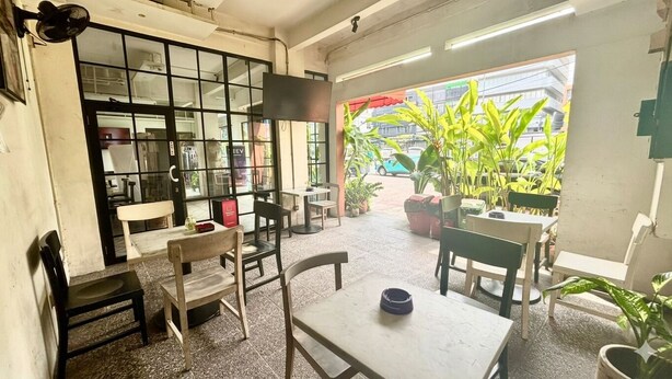 Un área de estar interior con mesas y sillas, un ventanal con vista a la calle, y plantas en macetas.