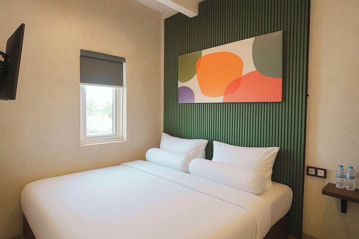 Ein Hotelzimmer mit einem großen Bett, einem Fenster mit Jalousien, einem an der Wand montierten Fernseher und einem farbenfrohen, abstrakten Gemälde.