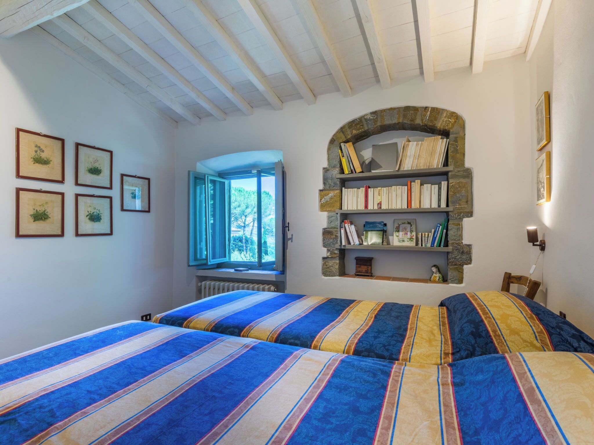 A bedroom with two beds, a stone bookshelf, framed pictures, and a window with an open shutter.