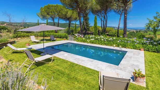 Une piscine rectangulaire bordée de pierres, entourée d’une pelouse bien entretenue et de diverses plantes. On y trouve deux chaises longues et un parasol de patio.