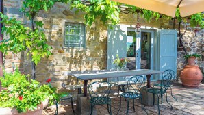Un bâtiment en pierre avec une table et des chaises placées sous un auvent, le tout entouré de verdure.