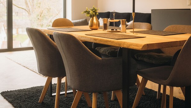 Un espace de salle à manger moderne avec une table en bois, des chaises à revêtement gris, un éclairage suspendu et une grande fenêtre qui laisse entrer la lumière naturelle.