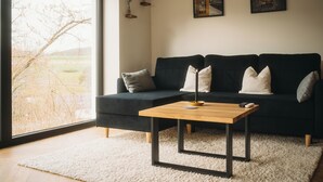 Un salon moderne avec un canapé foncé, une table basse en bois et une grande fenêtre donnant sur un arbre et un immeuble.