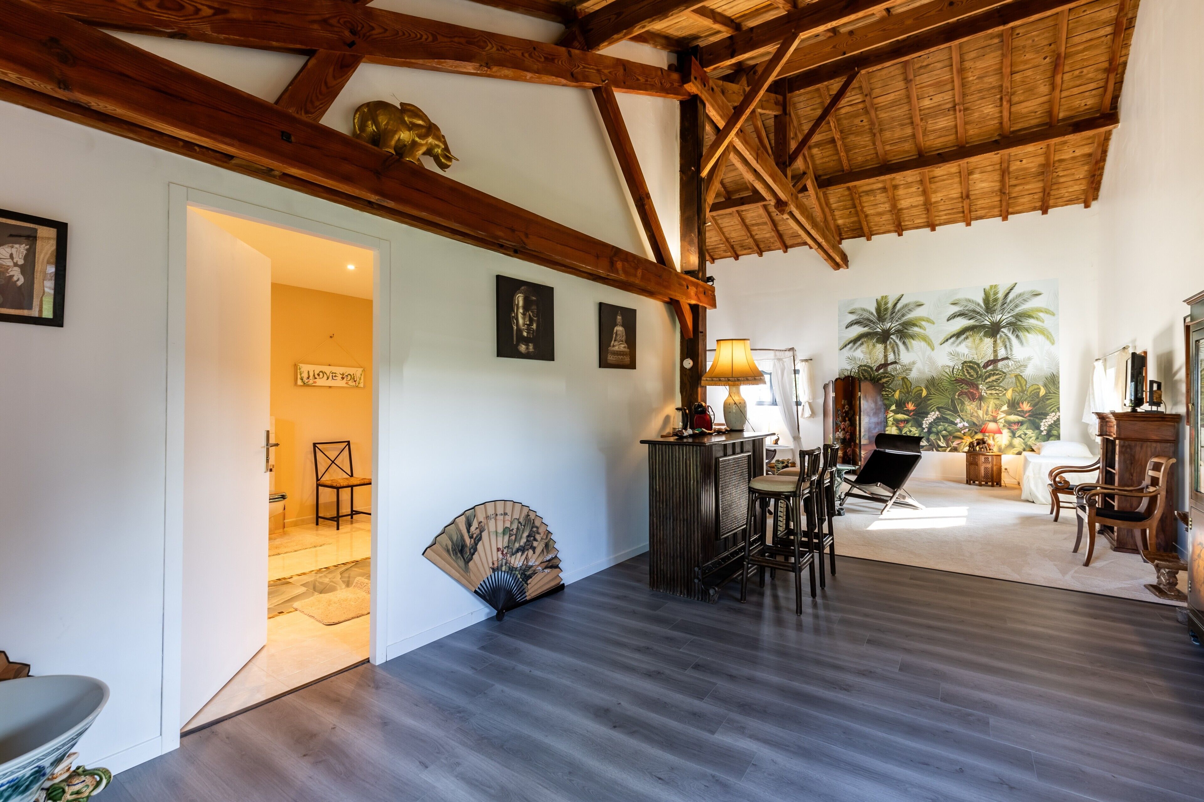 Une pièce spacieuse avec un plafond haut, des poutres en bois, un ventilateur, un coin bar et une vue sur des palmiers.