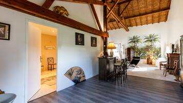 Une pièce spacieuse avec un plafond haut, des poutres en bois, un ventilateur, un coin bar et une vue sur des palmiers.