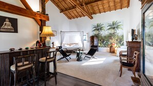 A spacious room with a high ceiling, a bed, a sitting area with chairs, a bar area, and a view of palm trees through a large window.