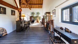 A spacious room with a wooden ceiling, a dining area with chairs, a bar area with a lamp, and a large window with a view of palm trees.