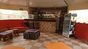 Un café avec un sol à carreaux, un comptoir où sont exposés divers articles, un réfrigérateur et un espace salon avec un canapé et des tables.