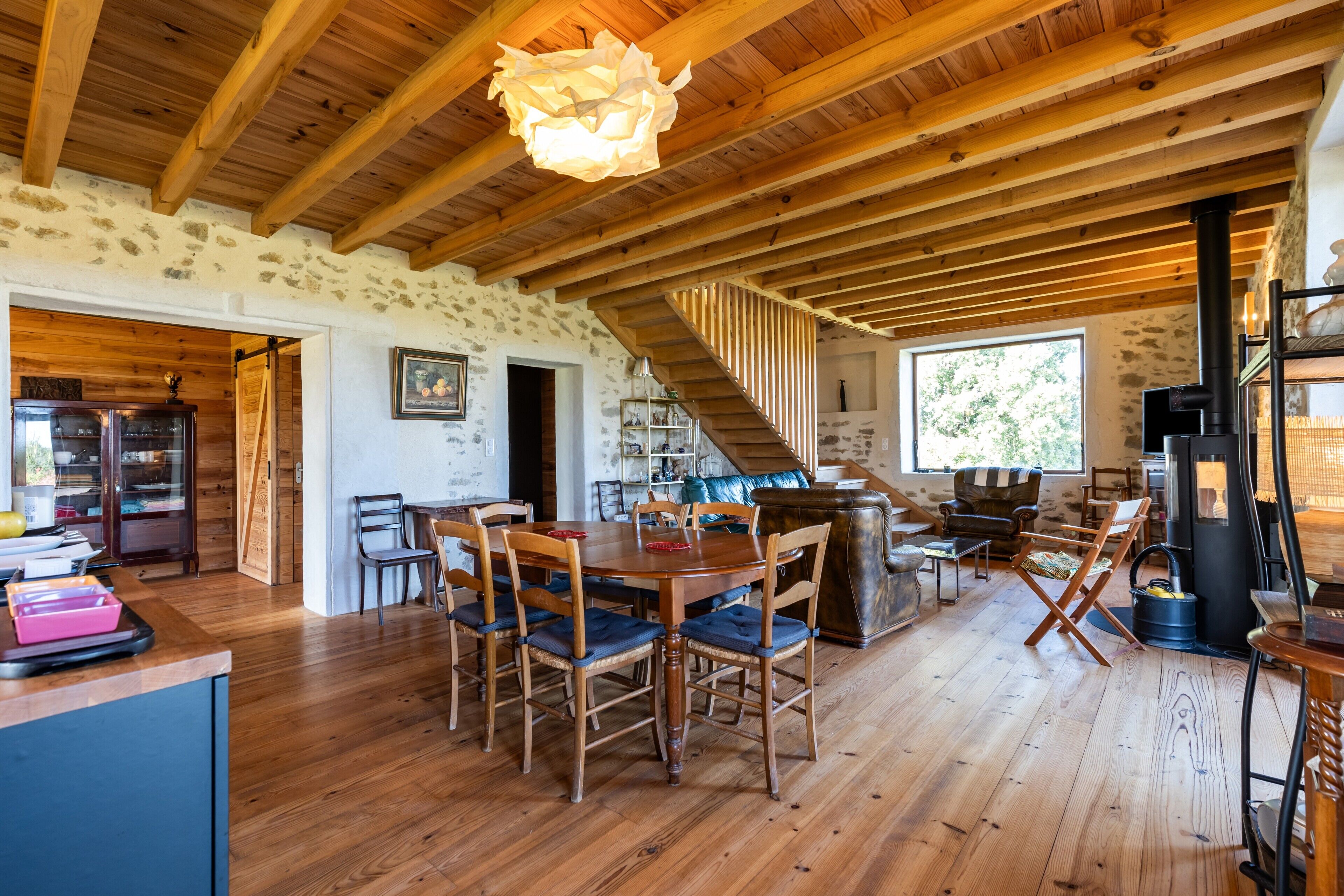 A rustic interior with a wooden dining table, chairs, a staircase, and a stone wall.