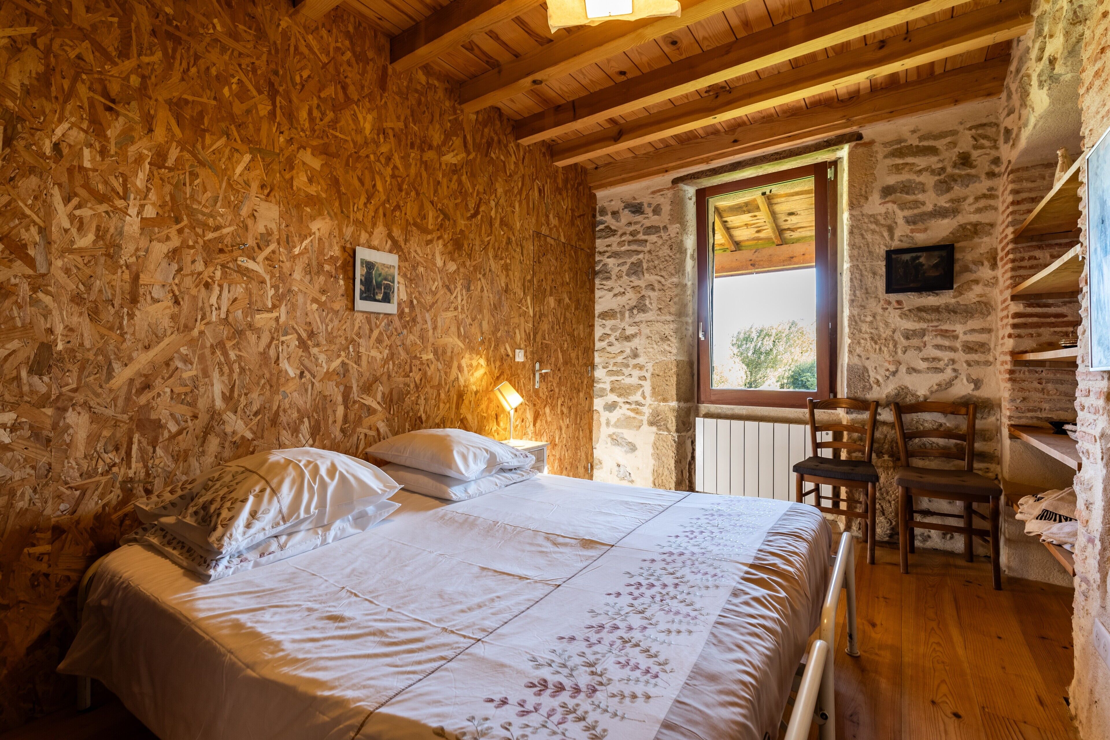 Una camera da letto con soffitto in legno, pareti in pietra, un letto con lenzuola a motivi floreali, un comodino, una sedia e una finestra con vista.