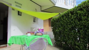 A patio with a table covered in a green tablecloth, a chair, and a basket with flowers. There is a white balcony railing and a hedge in the background. - Villa With Garden and Beach Place (San Michele al Tagliamento)