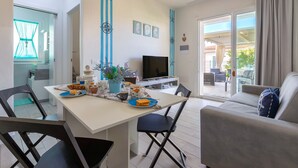 Una sala de estar moderna con sofá, mesa de comedor, sillas, televisor y balcón con mobiliario exterior.