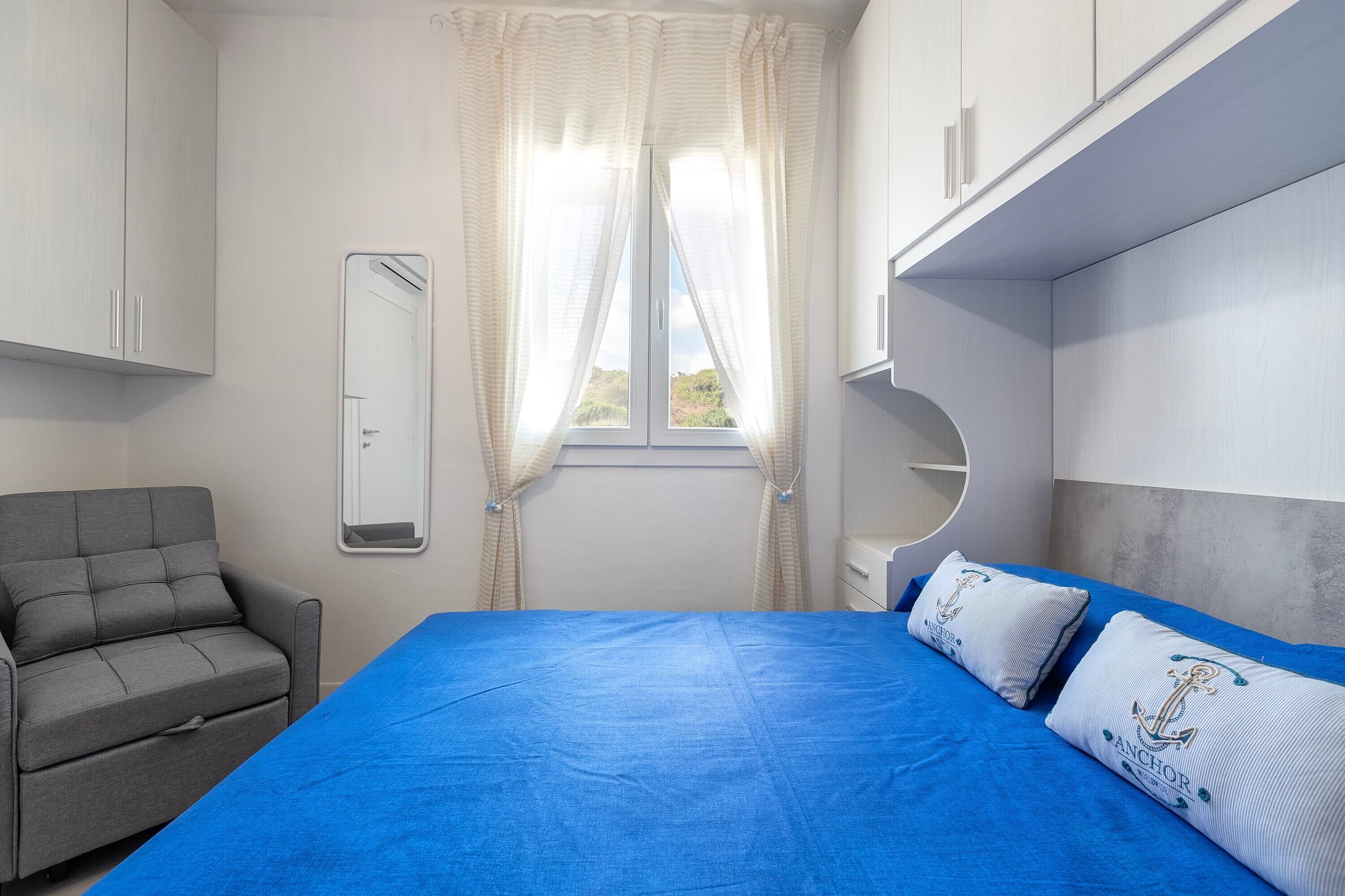 Un dormitorio con cama, un sofá gris, una ventana con cortinas y un espejo.