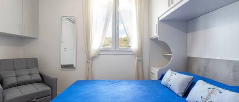 Un dormitorio con cama, un sofá gris, una ventana con cortinas y un espejo.