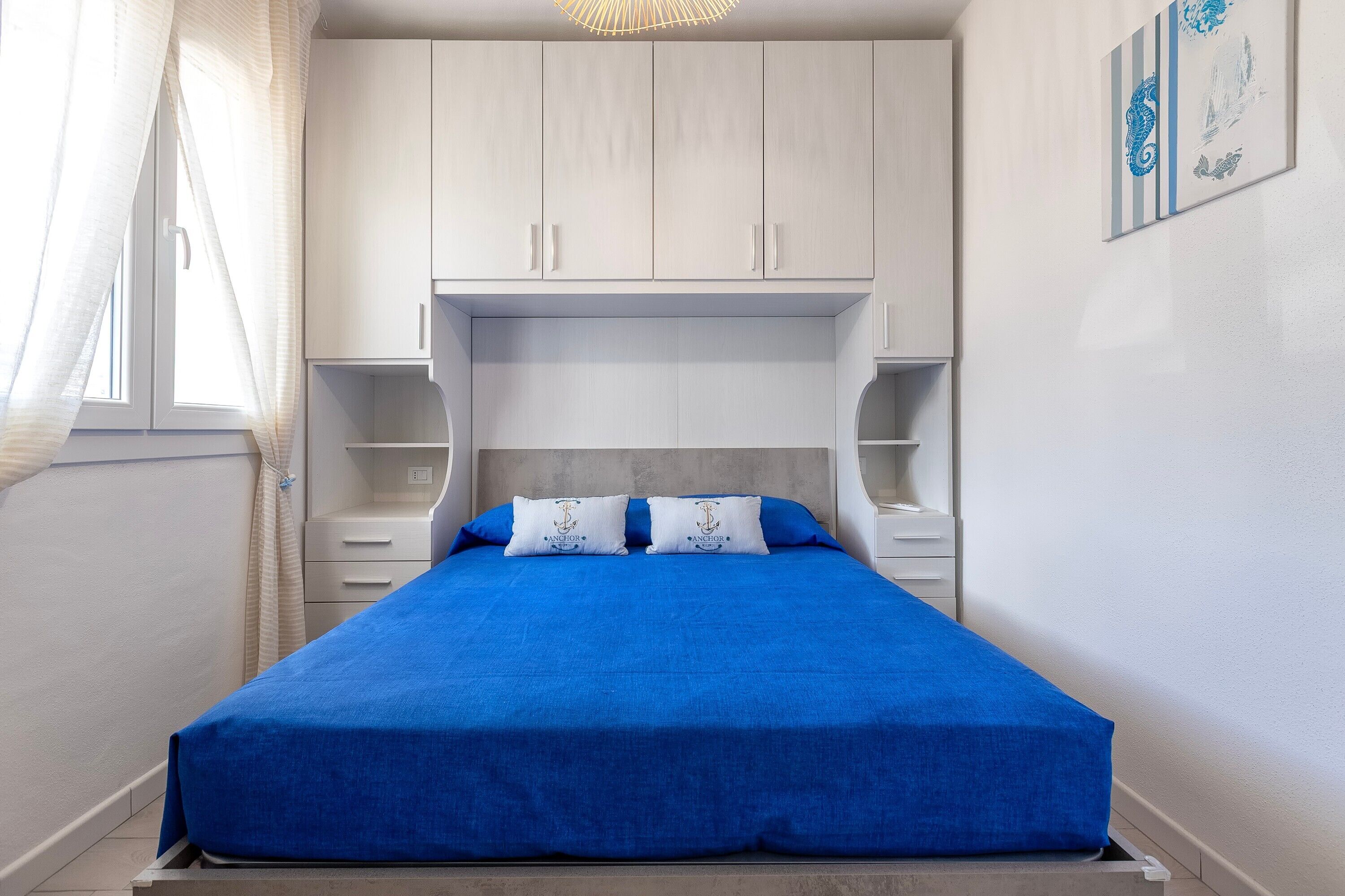 A bedroom with a bed, white cabinets, a window with curtains, and a wall-mounted artwork.