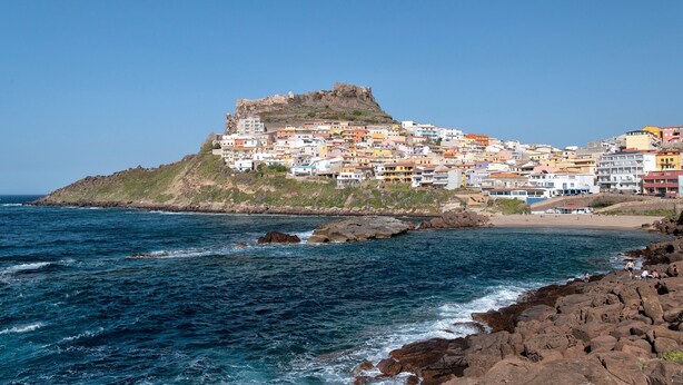 Un pueblo costero con edificios coloridos, un litoral rocoso y una colina destacada con una estructura similar a una fortaleza.