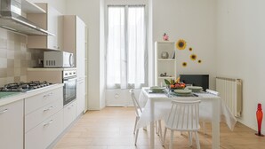 A modern kitchen with white cabinets, a gas stove, and a microwave. There is a dining table with chairs and a window with curtains. - The Houses on the Park 2 (La Spezia)