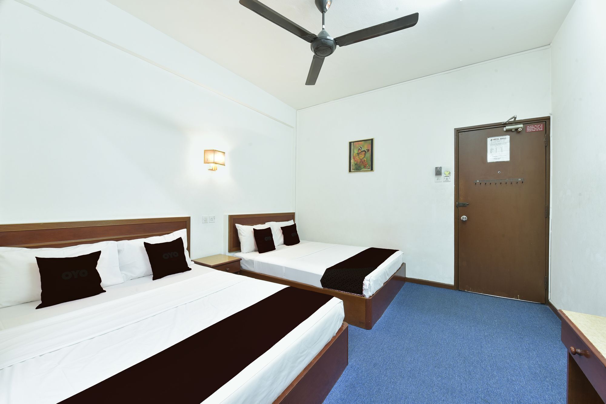 Habitación de hotel con dos camas, un ventilador de techo, una lámpara fijada a la pared, una puerta con un letrero y un pequeño cuadro en la pared.