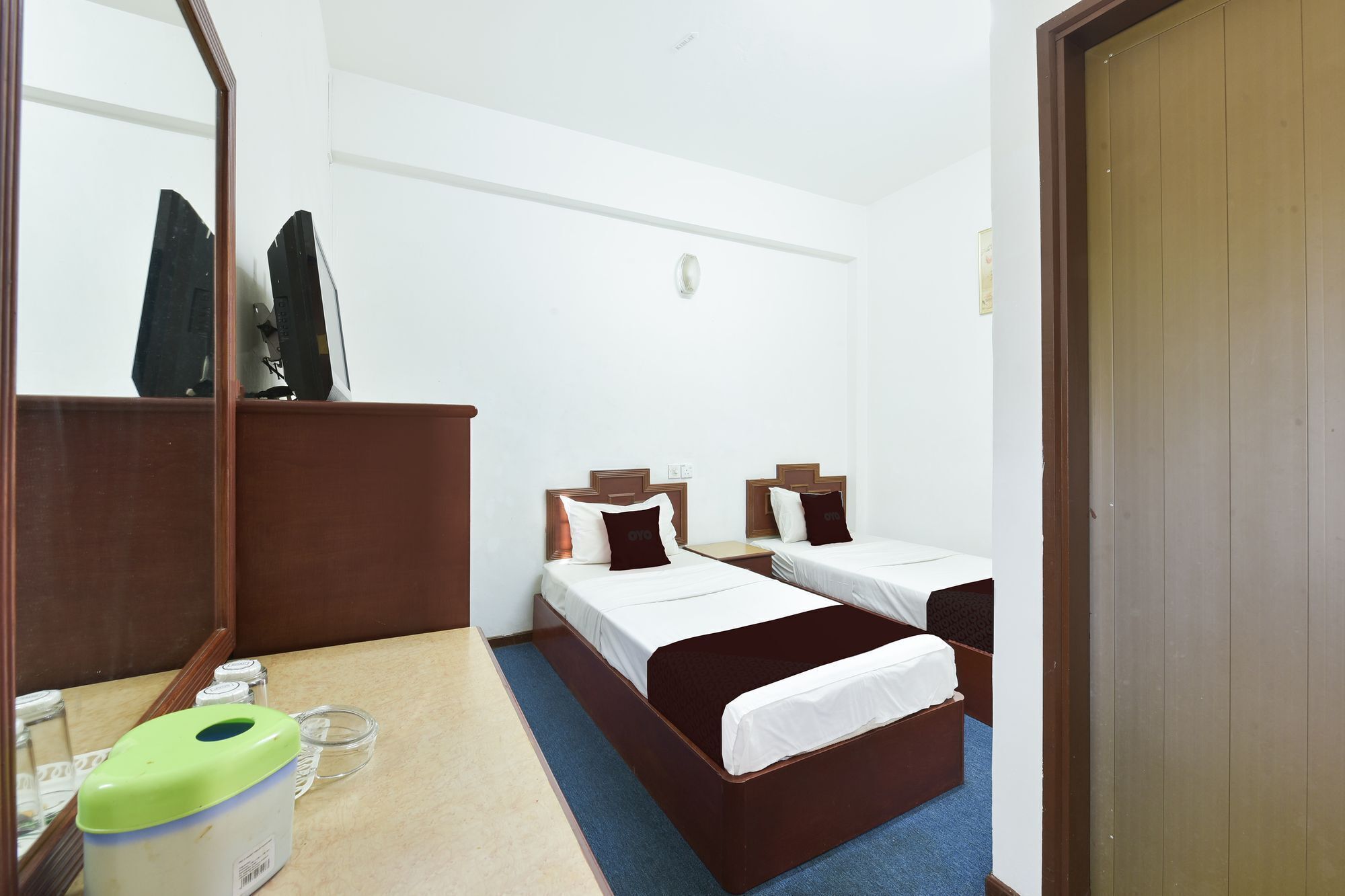 A hotel room with two single beds, a wooden wardrobe, a mirror, a small table with a tissue dispenser, and a blue carpet.