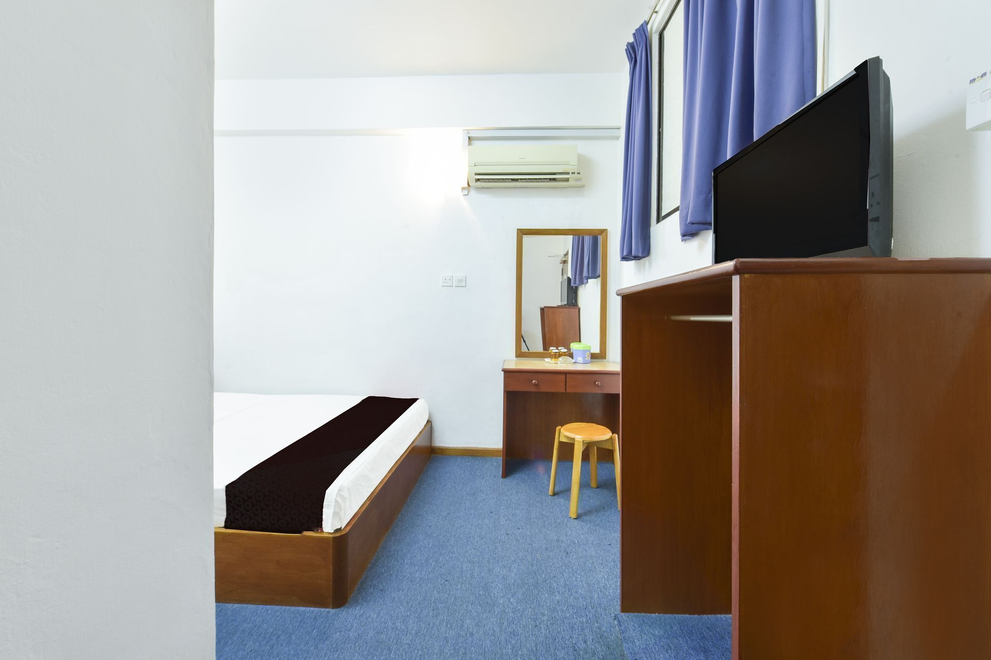 A compact hotel room with a single bed, a wooden desk, a chair, a mirror, a flat-screen TV, and an air conditioning unit.