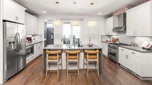 A modern kitchen with white cabinets, stainless steel appliances, a central island with bar stools, pendant lighting, and a view to the dining area.