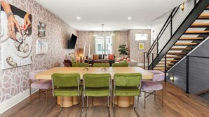 A modern dining area with a long table and green upholstered chairs, a staircase, and a living area with a sofa and a TV.