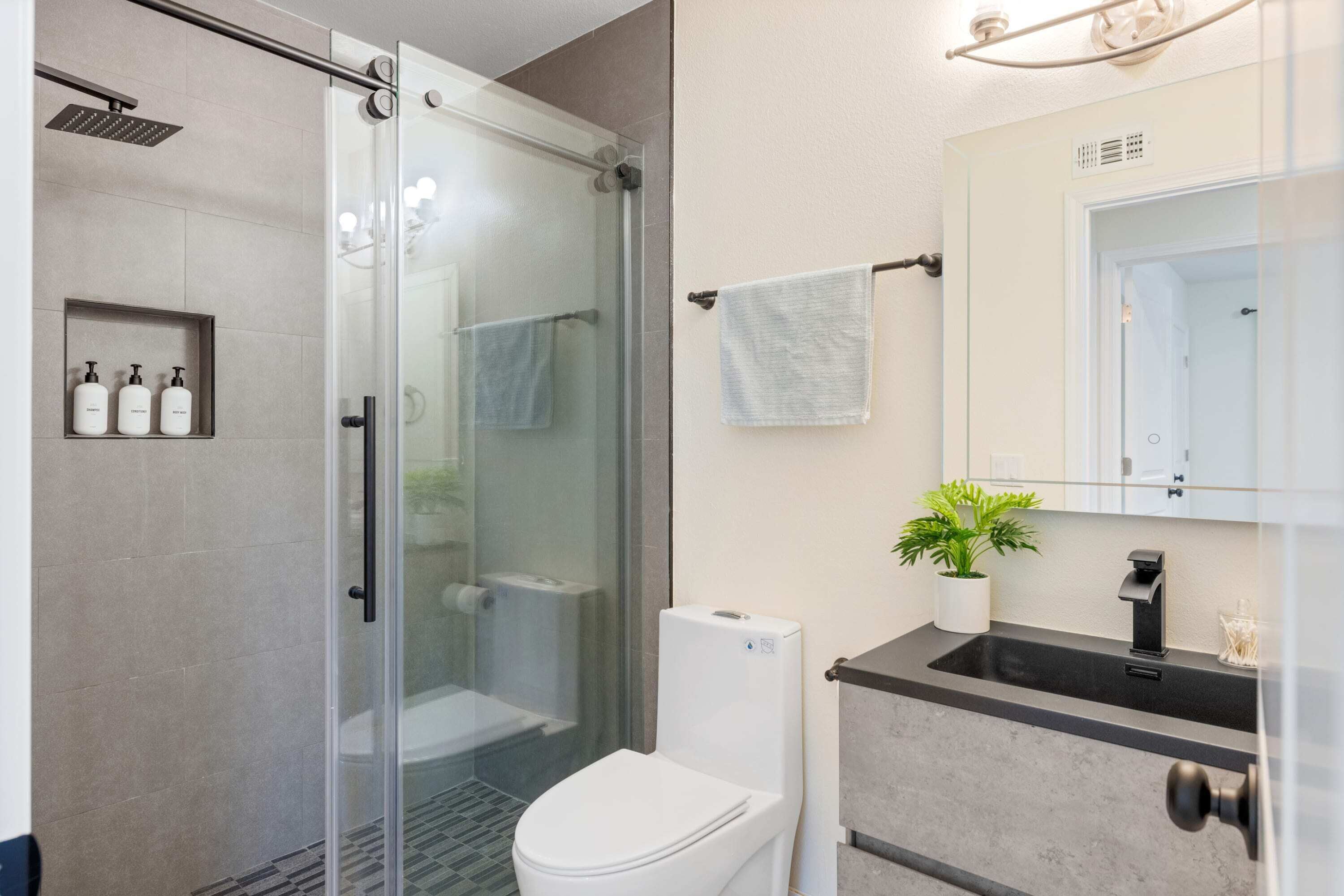 Une salle de bain moderne avec une douche à parois de verre, des toilettes blanches, un lavabo doté d’un plan de travail noir et un miroir au-dessus du lavabo.