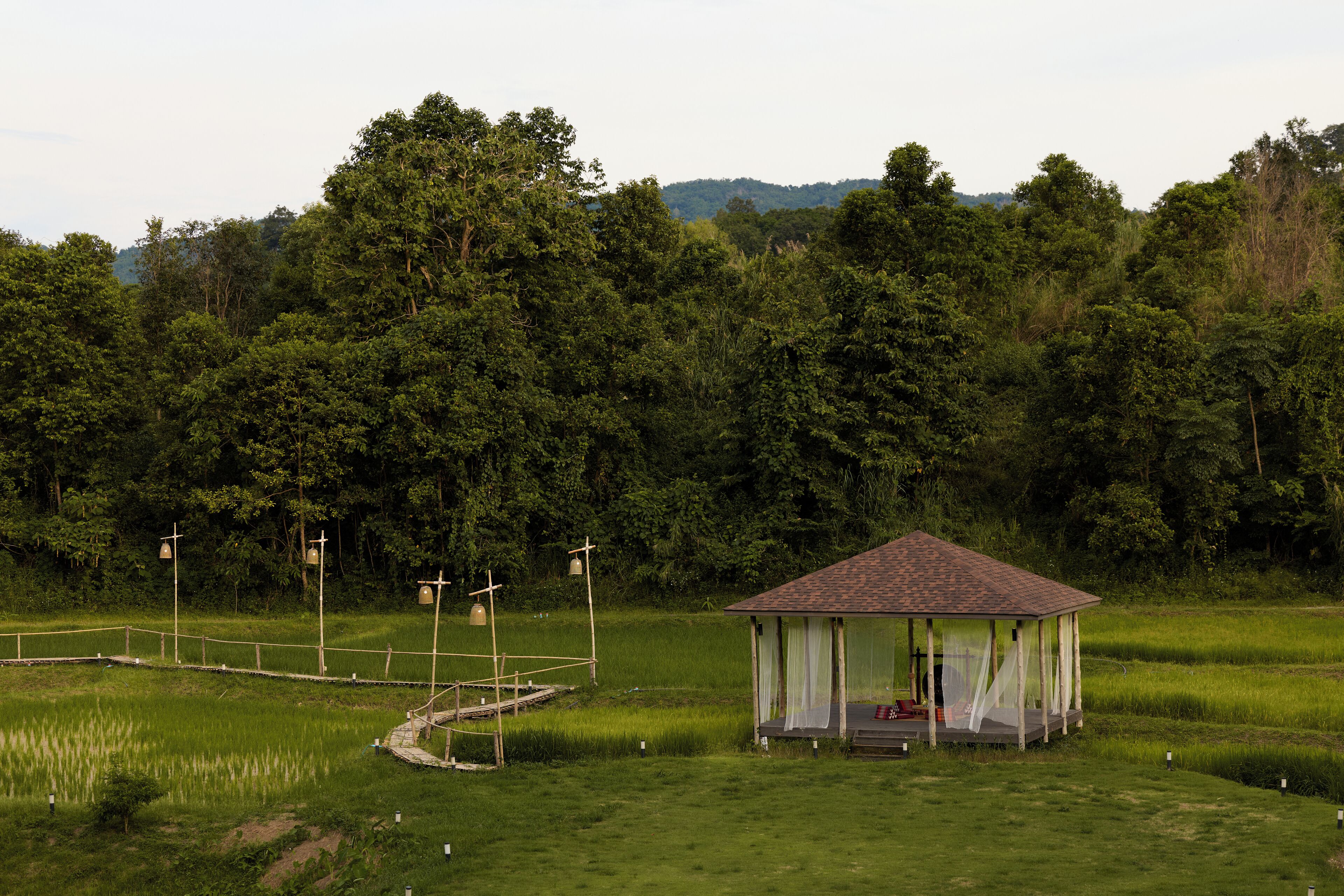 A gazebo with a tiled roof and a covered porch, surrounded by a grassy field and dense forest.