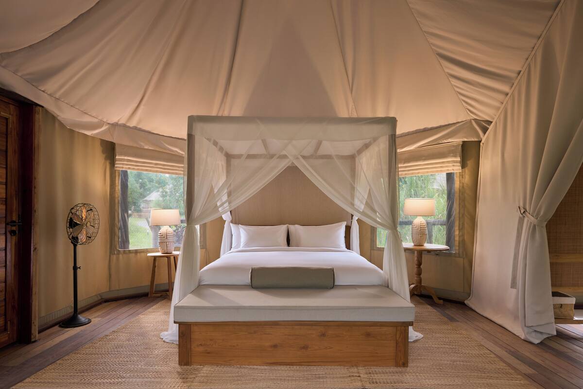 A four-poster canopy bed with white linens, two bedside tables with lamps, and a wooden floor.
