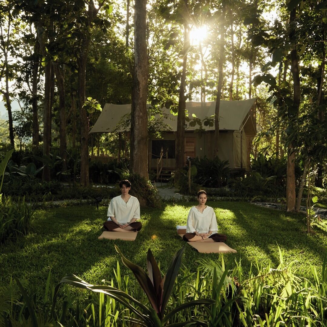 Two individuals sitting on mats in a lush garden with a house in the background.