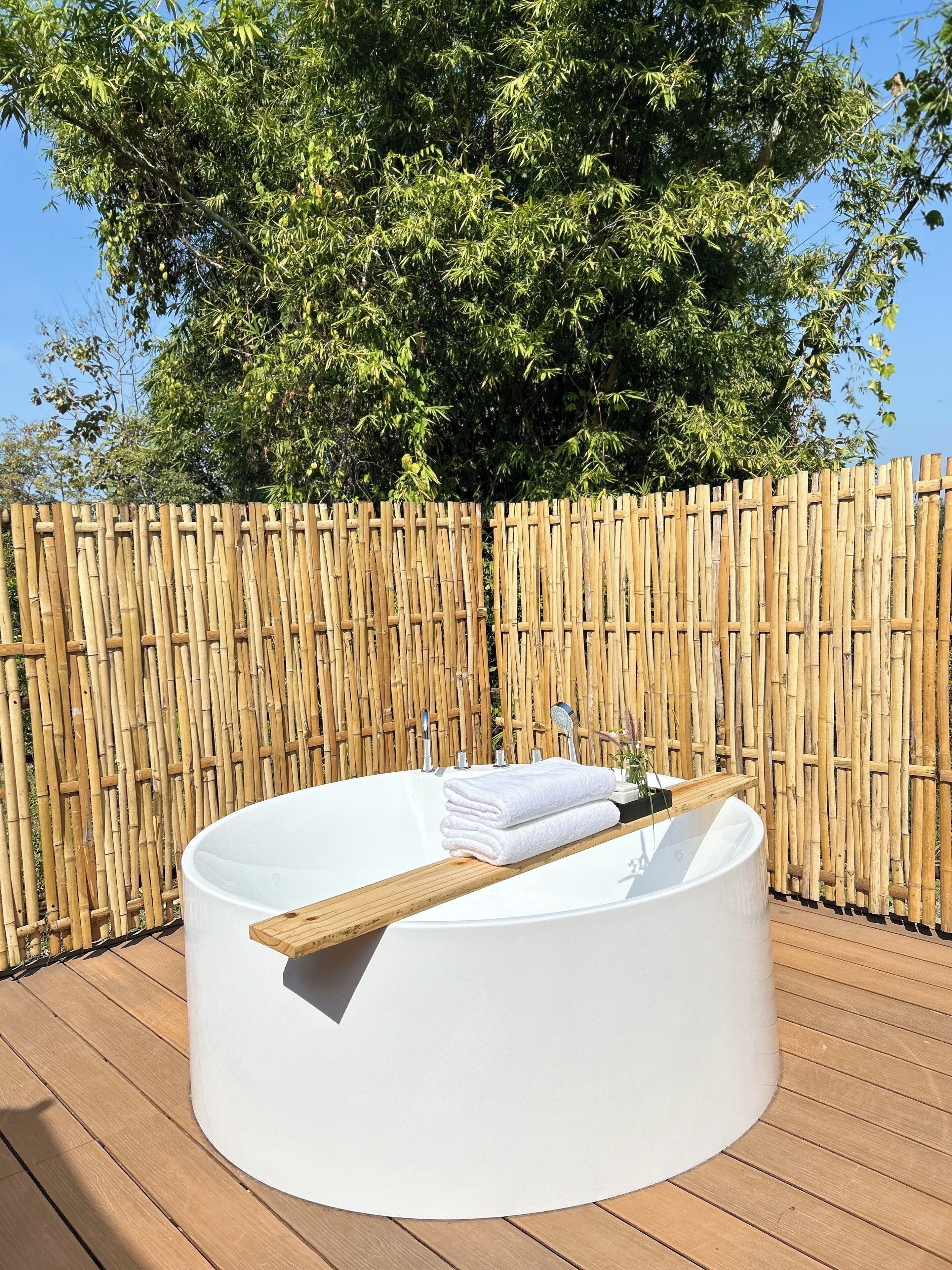 A round outdoor bathtub with a wooden tray and towels, surrounded by a bamboo fence and trees.