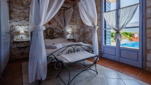 Une chambre avec un mur en pierre, un lit à baldaquin, un banc et une vue sur une piscine, visible à travers une porte.