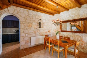 Comedor con paredes de piedra, una mesa y sillas de madera, una araña y una cocina visible a través de un arco.