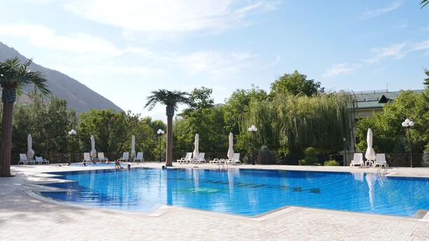 Una piscina rectangular con agua azul cristalina, rodeada de sillas de descanso blancas y sombrillas, con un fondo de vegetación exuberante y una montaña.