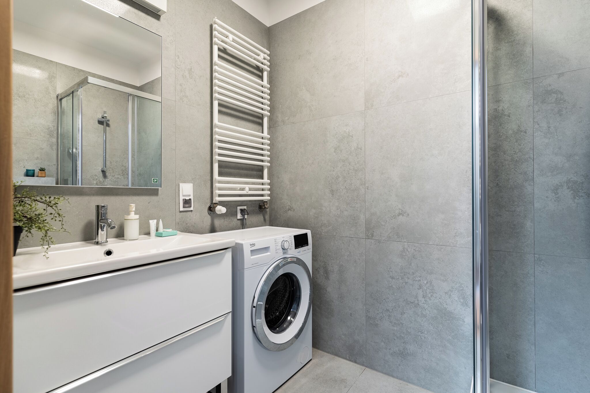 A modern bathroom with a built-in washing machine, a glass shower enclosure, and a large mirror.
