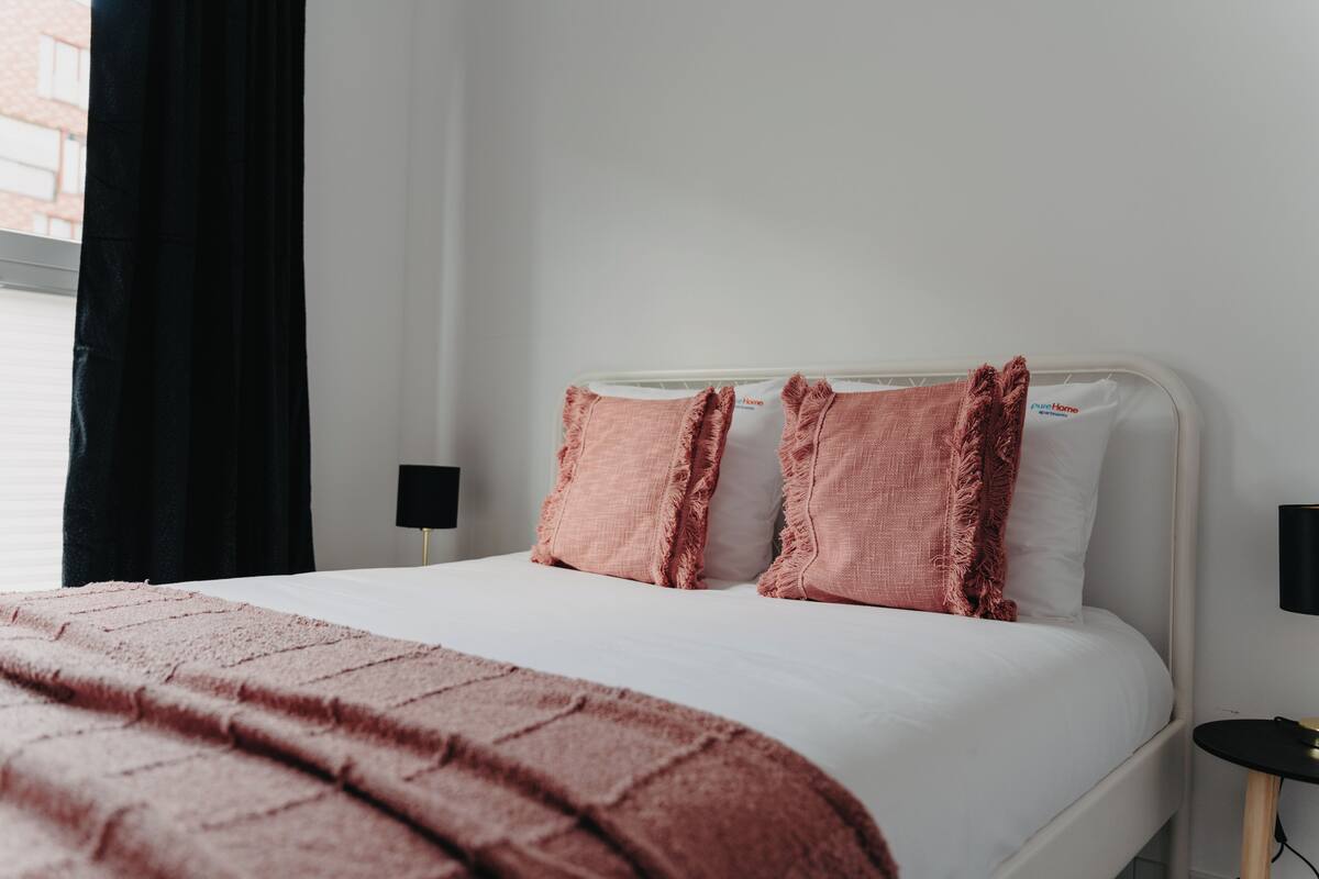 A neatly made bed with a white mattress and pink pillows, a pink blanket, and a black curtain partially drawn.